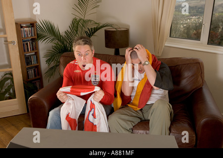 Parution du modèle. L'Angleterre et l'Allemagne de football fans assis sur le canapé à regarder la télévision que l'Angleterre gagne et perd de l'Allemagne. Banque D'Images