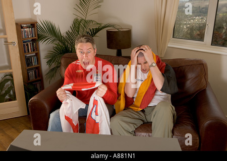 Parution du modèle. L'Angleterre et l'Allemagne de football fans assis sur le canapé à regarder la télévision que l'Angleterre gagne et perd de l'Allemagne. Banque D'Images