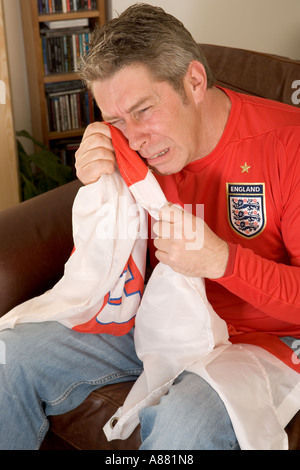 Angleterre football fan assis à la maison sur le canapé devant le plat de pleurer dans son St George's drapeau, son équipe vient de perdre. Banque D'Images