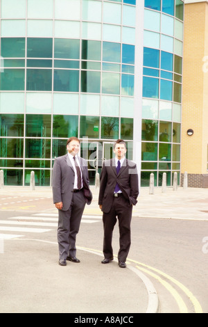 Deux hommes d'affaires caucasiens en costumes debout à l'extérieur d'un immeuble de bureaux moderne avec une façade vitrée courbée Banque D'Images