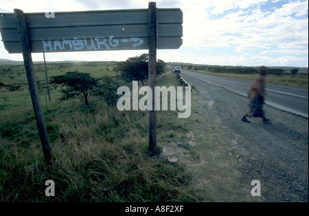 Une femme traverse une route goudronnée derrière un panneau peint à la main à Hambourg dans l'Eastern Cape, Afrique du Sud. Banque D'Images