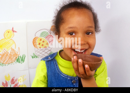Heureux bébé fille Afro Caraïbes mordre dans une coquille d'œufs de Pâques au chocolat Banque D'Images