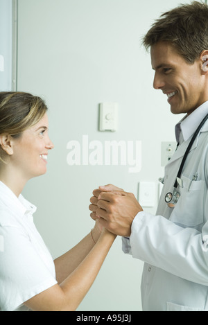 Médecin et femme debout face à face, holding hands and smiling at each other Banque D'Images