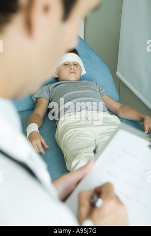 Garçon couché sur la table d'examen avec bandage sur le front et le bras, doctor writing on clipboard Banque D'Images