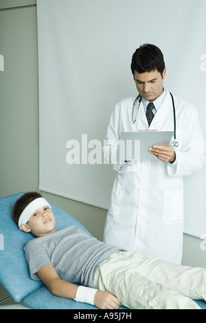 Garçon couché sur la table d'examen avec bandage sur le front et le bras, doctor writing on clipboard Banque D'Images