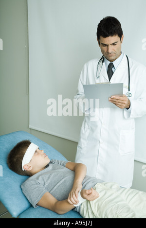 Garçon couché sur la table d'examen avec bandage sur le front, tenant le bras, doctor writing on clipboard Banque D'Images