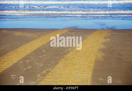 Un jeu de pneu ou de chenilles traversant une plage de sable clair et disparaître dans ou sortant d'un doux bleu de la mer Banque D'Images