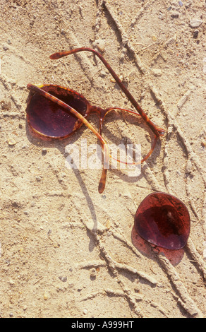 Close up of brisé et paire de lunettes de soleil avec une seule lentille allongé dans le sable lâche avec traces d'exécution sous l'emprise et Banque D'Images