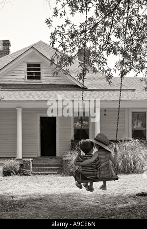 Frère et soeur avec leurs bras autour de l'autre se balançant sur une ancienne tree swing en face d'une ferme restaurée. Banque D'Images