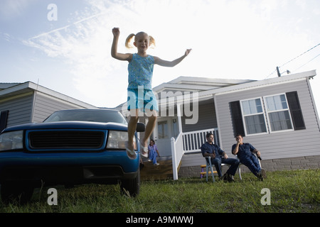 Girl Jumping off pare-choc de vieux camion garé en face de la maison, famille en arrière-plan Banque D'Images