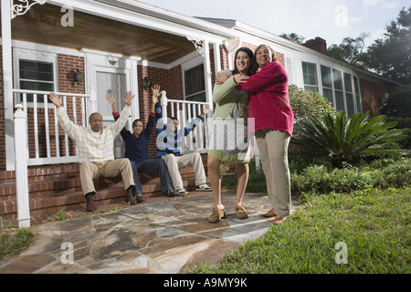 Woman embracing sa belle-fille, en face de la maison avec la famille en arrière-plan Banque D'Images