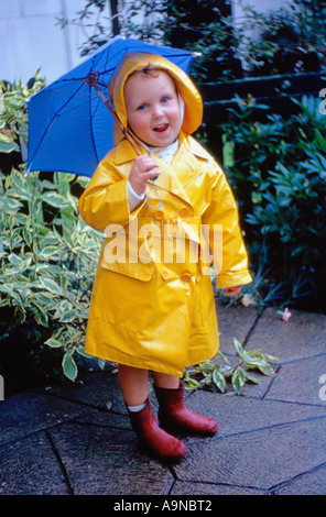 Petit enfant dans un imperméable jaune vif et un chapeau sou'wester, tenant un parapluie bleu, dans des bottes rouges en wellie, dans un jardin britannique. Vers 1960 Banque D'Images