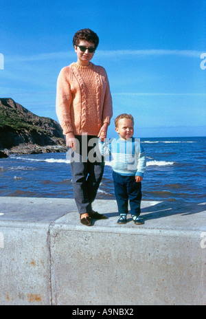 Jeune femme caucasienne et petit enfant en pulls tricotés, debout main dans la main sur un mur de front de mer britannique, années 1960 Banque D'Images