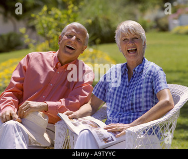 L'âge à la retraite couple assis sur les chaises en osier dans le jardin et de rire à l'appareil photo Banque D'Images