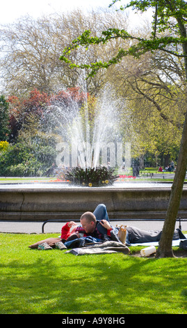 Deux amoureux allongés sur l'herbe dans un parc public avec une fontaine jaillissante en arrière-plan Banque D'Images