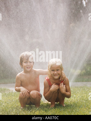 Petit garçon et fille jouant dans sprinkleur pelouse Banque D'Images