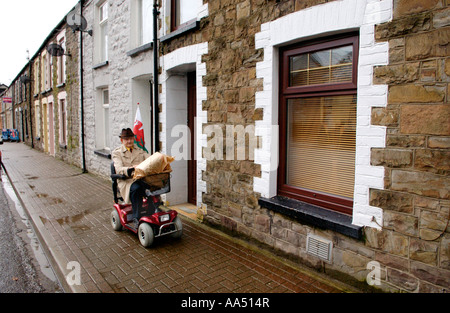 Vieil homme sur la mobilité scooter flying dragon rouge d'un drapeau tandis que dehors sur la rue commerçante de Treorchy, Valley South Wales UK Banque D'Images