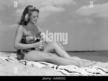 1950 FEMME EN UNE SEULE PIÈCE SANS BRETELLES MAILLOT assis sur une serviette de plage à mettre Lait Solaire Banque D'Images