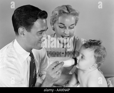 1950 SMILING PÈRE nourrir bébé LAIT DE BOUTEILLE AVEC MÈRE À SIDE Banque D'Images