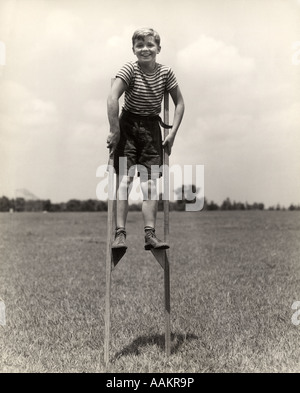 Années 1930 Années 1940 SMILING HAPPY BOY WEARING STRIPED SHIRT & pantalon court MARCHE SUR PAIRE DE PILOTIS LOOKING AT CAMERA Banque D'Images