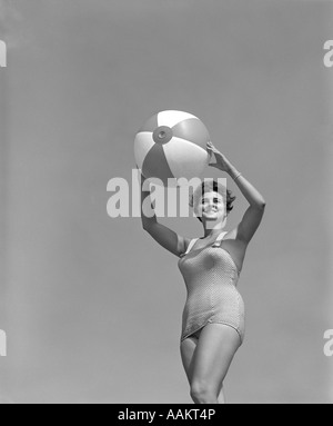 1950 SMILING WOMAN EN UN SEUL MORCEAU MAILLOT HOLDING BEACH BALL AU-DESSUS DE LA TÊTE À LA Banque D'Images