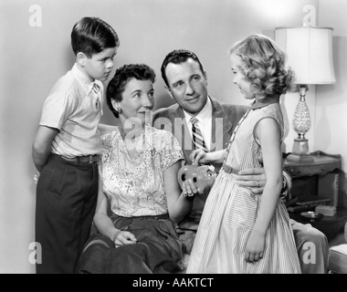 Années 1950 LA FAMILLE DE LA MÈRE HOLDING PIGGY BANK FILLE TOMBER DANS COIN PÈRE ET FILS À LA SUR Banque D'Images