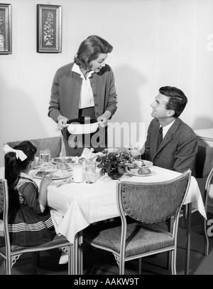 Années 1950 Années 1960 FAMILLE DE TROIS À Table de salle à dîner À PARLER AVEC FEMME DEBOUT METTRE COCOTTE SUR TABLE Banque D'Images