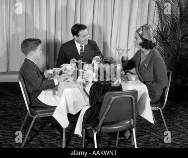 1960 SMILING HAPPY FAMILLE DE MANGER QUATRE REPAS AU RESTAURANT PAPA PARLER À MAMAN QUI PORTE STYLE TURBAN HAT Banque D'Images