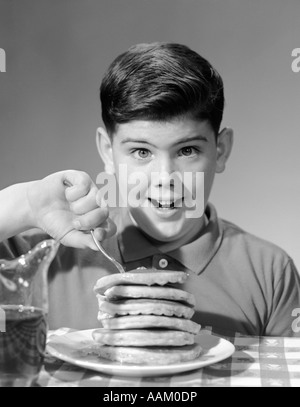 1960 SMILING PORTRAIT garçon assis derrière avec pile de crêpes épaisses DE PIQUER LA FOURCHE DANS LE HAUT DE LA PILE LOOKING AT CAMERA Banque D'Images