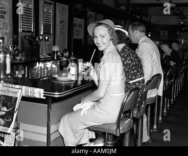 1940 SMILING WOMEN habillé en robe de lin HAT HOLDING GLASS COLA ASSIS AU COMPTOIR SODA FOUNTAIN LOOKING AT CAMERA Banque D'Images