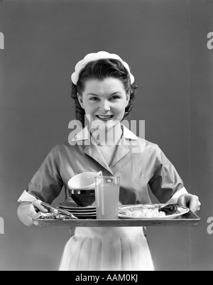 1940 SMILING WAITRESS PORTANT UNE CASQUETTE ET UNIFORME AVEC TABLIER & COL BLANC TENANT UN PLATEAU DE SERVICE LOOKING AT CAMERA Banque D'Images
