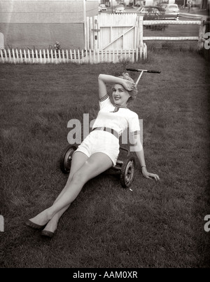 1950 ÉPUISÉ WOMAN WEARING WHITE MINI SHORTS SITUÉE AU-DESSUS D'UNE TONDEUSE À POUSSER LOOKING AT CAMERA Banque D'Images