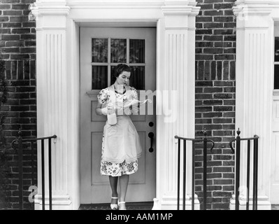 1940 FEMME DANS UN tablier, debout à l'extérieur de la porte tenant une pinte de lait et READING NEWSPAPER Banque D'Images