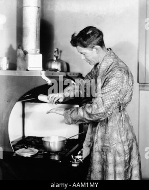 Années 1920 Père habillé en robe DEBOUT À POÊLE TESTER LA TEMPÉRATURE DU LAIT SUR SA MAIN D'UN BIBERON CHAUD Banque D'Images