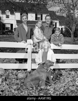 1960 Portrait de famille Père Mère FILS ET DEUX FILLES CHIEN RÉUNIS AUTOUR DE COUR AVANT WHITE FENCE LOOKING AT CAMERA Banque D'Images