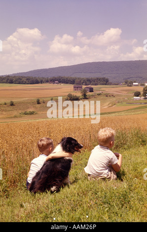 1960 RETOUR DE DEUX GARÇONS AVEC LE NOIR ET BLANC CHIEN ASSIS SUR LE TERRAIN À flanc de colline jusqu'à la ferme Banque D'Images