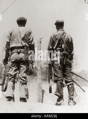 Années 1930 Années 1940 DEUX HOMMES RETOUR VUE ARRIÈRE POTEAU DE TRAVAILLEURS PORTANT CEINTURE CHARGÉE DE L'ÉQUIPEMENT ET DES OUTILS Banque D'Images