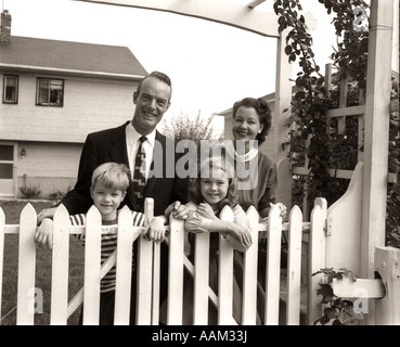 1950 FAMILLE DE QUATRE DERRIÈRE PICKET FENCE IN BACKYARD SMILING AT CAMERA Banque D'Images