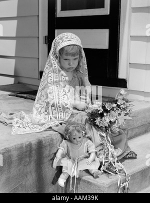 1950 GIRL IN MOCK TOGE AVEC BOUQUET À genoux et bébé poupée à pieds avec regard triste SUR LE VISAGE Banque D'Images