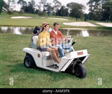 1960 L'HOMME ET LA FEMME DANS LA CONDUITE DE CHARIOT DE GOLF À TRAVERS L'HERBE PRÈS DE L'EAU DE VIE PASSE-TEMPS DEUX CLUBS DE GOLF BAG Banque D'Images