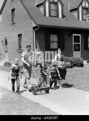 1950 FAMILLE DE CINQ SUR TROTTOIR DE BANLIEUE À MARCHER AVEC TONDEUSE Outils de jardinage et fleurs Banque D'Images