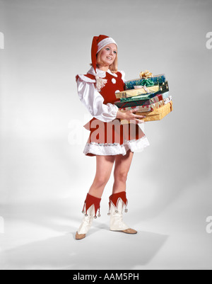 1960 YOUNG WOMAN IN CHRISTMAS SANTA HELPER COSTUME ROUGE ET BLANC ET CAP HOLDING PILE OF GIFT WRAPPED PRESENTS Banque D'Images