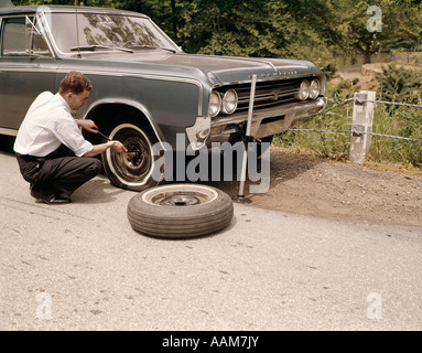 1960 MAN CHANGING CREVAISON SUR VOITURE À CÔTÉ DU CHEMIN RURAL OUTILS CRIC DE VOITURE À CHANGER LES PNEUS Banque D'Images