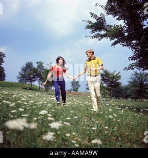 1970 YOUNG TEEN COUPLE HOLDING HANDS RUNNING THROUGH FIELD QUEEN ANNE'S LACE Fleurs sauvages ROMANCE FUN FASHION Banque D'Images