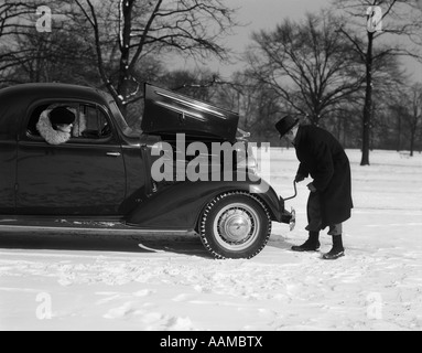 Années 1930 Années 1940 Femme regardant PASSAGER AUTOMOBILISTE HOMME ESSAYER DE LA MANIVELLE D'UN CHEVROLET COUPé CALÉ DANS LA NEIGE Banque D'Images