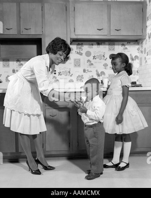 1960 SMILING AFRICAN AMERICAN WOMAN MOTHER IN APRON ET POMPES DONNANT UN VERRE DE LAIT À SON FILS ET SA FILLE AVEC MARY JANE SHOES Banque D'Images