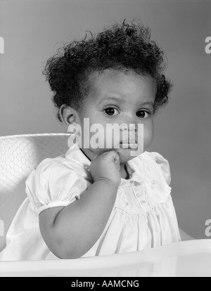 1960 AFRICAN AMERICAN BABY GIRL IN DRESS SITTING IN HIGH CHAIR WITH HAND ON CHIN & EXPRESSION GRAVE Banque D'Images