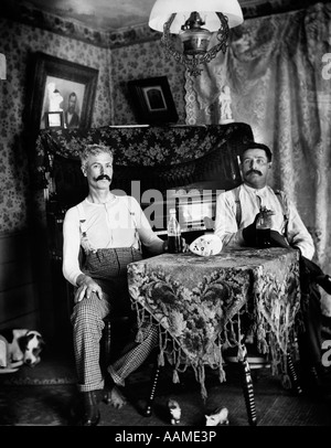 Années 1890, DEUX HOMMES EN CHEMISE ASSIS À TABLE BOISSONS EN BOUTEILLES DE BIÈRE AVEC PIANO ET LE CHIEN EN ARRIÈRE-PLAN Banque D'Images