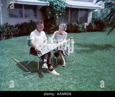 1950 MIDDLE AGED COUPLE assis dans des chaises de jardin en RÂTEAU SUR HERBE REGARDER LA CARTE DE VOYAGE TROPICAL RETRAITE Banque D'Images