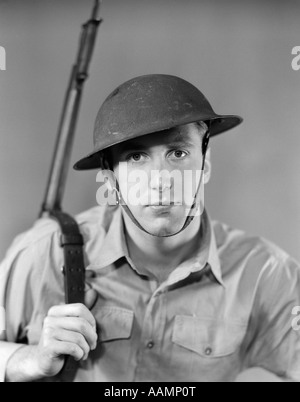 1940 PORTRAIT DE L'HOMME AMÉRICAIN SÉRIEUX SOLDAT FUSIL FUSIL D'EXPRESSION SUR L'ÉPAULE AVEC JUGULAIRE CASQUE WW2 ARMY VINTAGE Banque D'Images